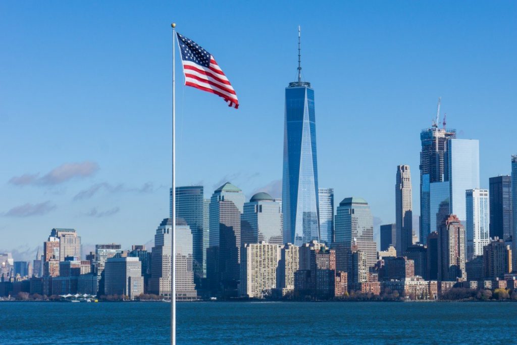 Americká vlajka, v pozadí řeka Hudson River a budova One World Trade Center.